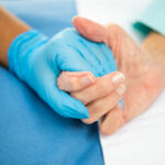 Close of Praise’s gloved hands holding what appears to be the hands of a patient. Caption
